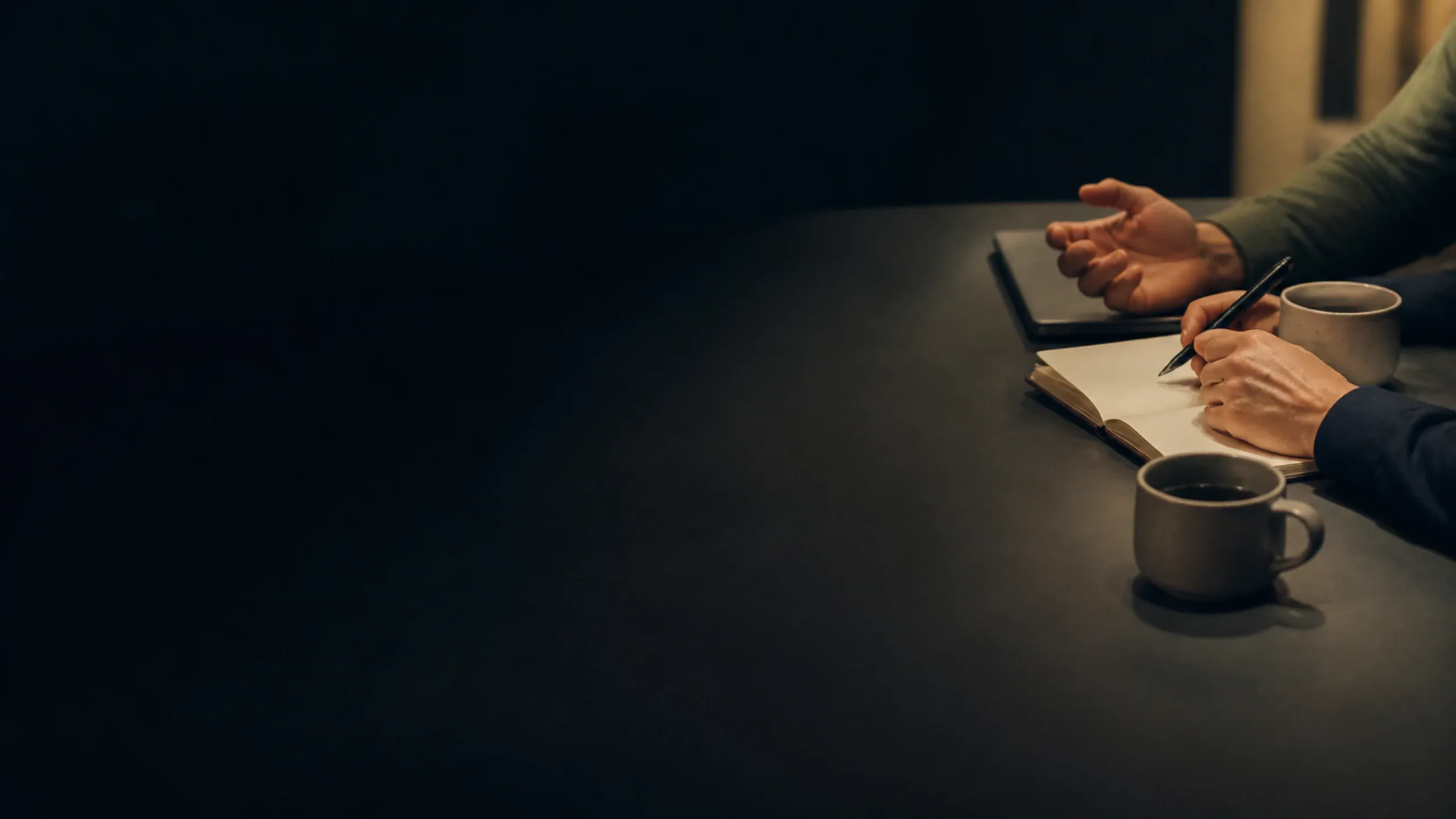 Twee handen schrijven in een notitieboek op een tafel, met een kopje koffie ernaast.